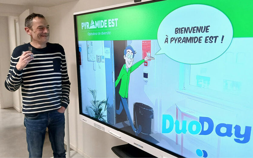 Un chargé de mission devant une grande télévision qui affiche le logo du DUO DAY et un petit personnage dessiné qui franchit la porte de PYRAMIDE EST en souhaitant la bienvenue à tout le monde.