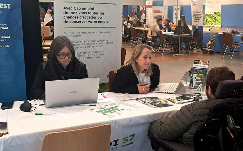 Deux chargées de mission sur un stand, l'une travaille sur son ordinateur et l'autre discute avec une personne en fauteuil roulant.