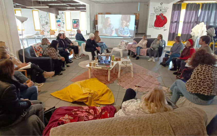 Une groupe de 20 personnes est réuni sur des poufs, des canapés et fauteuil et regardent un écran.